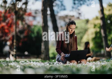 Young woman student working on laptop outdoors in park with headphones and focused expression Stock Photo