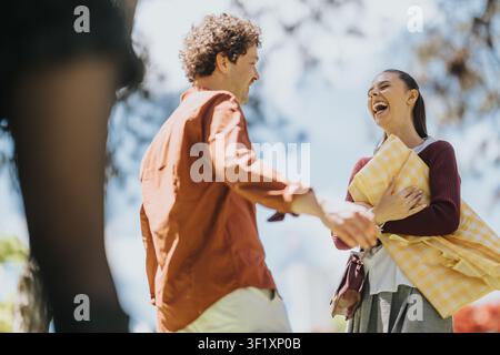 Young couple laughing and greeting outdoors while woman holds a yellow picnic blanket on a sunny day Stock Photo