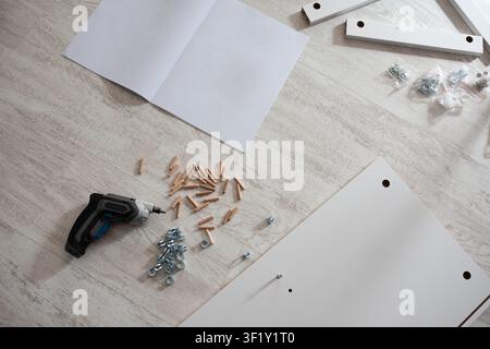Flat lay of furniture assembly process with wooden parts, metal screws, a cordless screwdriver and assembly instructions on a wooden light floor. Stock Photo