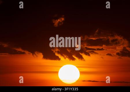 The glowing sun descends through clouds against a vivid red sky Stock Photo