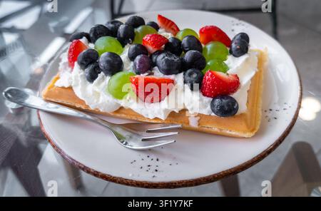 Waffle topped with whipped cream, fresh blueberries, green grapes, and strawberries on a white plate, ready to eat Stock Photo