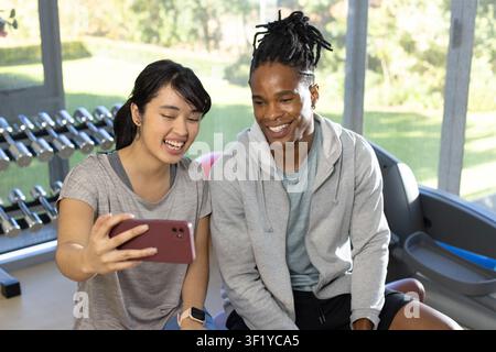 Diverse couple in athletic wear taking selfie on bench at gym with smartphone, dumbbell rack Stock Photo