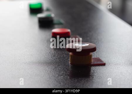 Close-up of a red emergency stop button mounted on a textured industrial control panel, with a blurred row of green and red indicator buttons receding Stock Photo