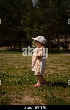 toddler girl in a pastel-striped sundress and white-pink cap standing in a sunlit grassy park, watching iridescent soap bubbles float by against dark Stock Photo