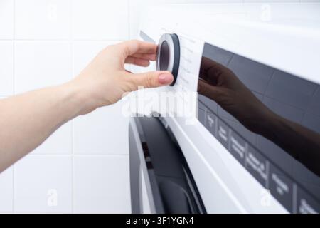 Close-up of a woman adjusting the program selector on a modern front-loading washing machine. Ideal for illustrating laundry routines, home appliances Stock Photo