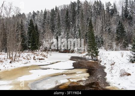 Partially frozen winter river winding through snow covered forest with dark flowing water and melting ice, natural Nordic landscape, Stock Photo
