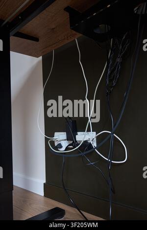 Tangled cables, power adapters and plugs connected to wall outlet under wooden desk. Concept of poor cable management and workspace organization Stock Photo