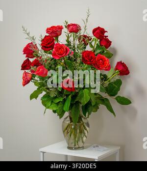 A lush bouquet of red and coral roses with greenery arranged in a clear glass vase on a white table. Stock Photo