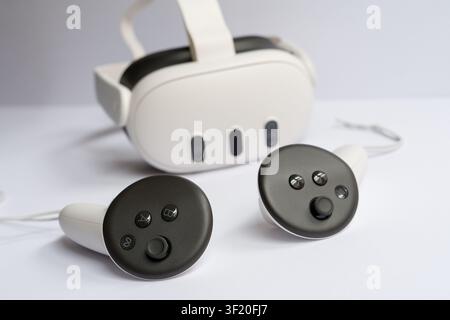 A close-up horizontal studio shot of a modern white VR (Virtual Reality) headset with two wireless hand controllers lying next to it on a clean white Stock Photo