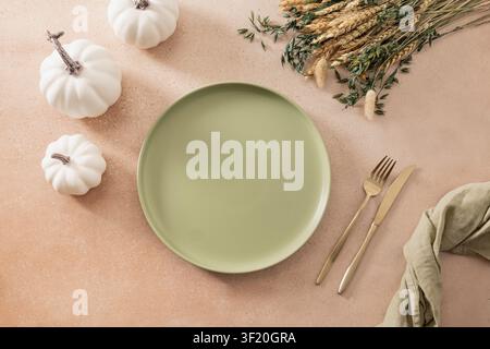 Elegant autumn table setting with green ceramic plate, white pumpkins, dried wheat and linen napkin on beige background. Minimal seasonal dining conce Stock Photo