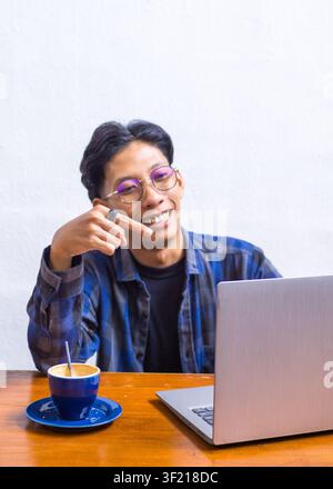 Cheerful young Asian man smiling enthusiastically while working on a laptop at a coffee shop, candid lifestyle portrait. Stock Photo