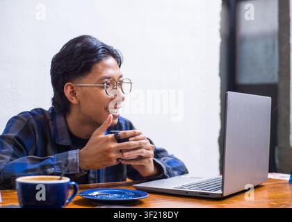 Cheerful young Asian man smiling enthusiastically while working on a laptop at a coffee shop, candid lifestyle portrait. Stock Photo