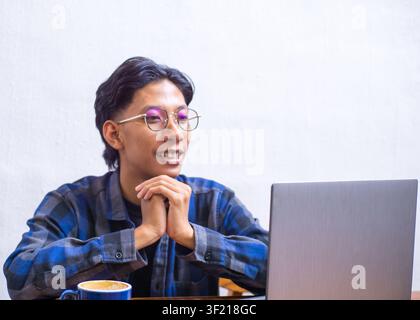 Cheerful young Asian man smiling enthusiastically while working on a laptop at a coffee shop, candid lifestyle portrait. Stock Photo