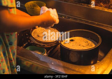 A close-up shot focused on a pile of unwashed plates and bowls covered in thick soap suds, with hands washing them in a kitchen sink. Stock Photo