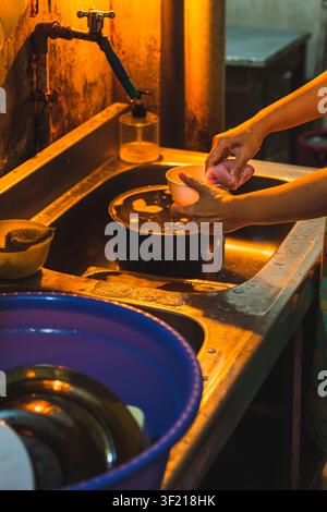 A person's hands scrubbing a small plastic bowl with a pink sponge over a stainless steel kitchen sink filled with soapy water. Stock Photo