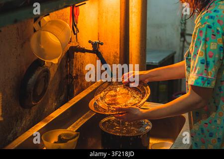 A woman wearing a patterned pajama shirt stands at a stainless steel kitchen sink, rinsing a clear glass plate under a running water faucet. Stock Photo
