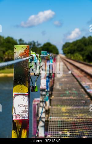 A weathered railroad trestle bridge covered in colorful graffiti and padlocks stretches over a calm river under a bright blue sky. Stock Photo
