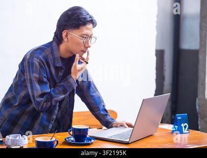 Focused young Asian man concentrating on his laptop while working remotely at a coffee shop, lifestyle portrait. Stock Photo