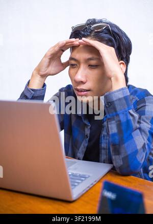 Stressed young Asian man holding his head in confusion while working on a laptop at a cafe, candid lifestyle concept portrait. Stock Photo