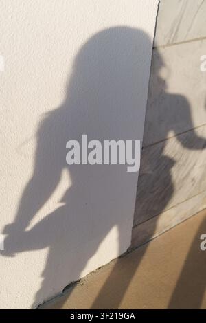 The elongated shadow of a person holding a camera stretches across a white plaster wall meeting a marble tiled surface in sunlight. Stock Photo