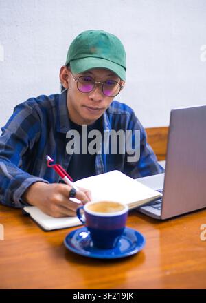 A young Asian man working on his laptop at a coffee shop, with close-ups of a blue ceramic coffee cup, notebook with a pen, and details of a remote wo Stock Photo
