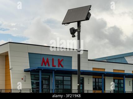 A solar panel and security camera mounted on a pole stand outside a modern school building with a red MLK sign under cloudy skies. Stock Photo