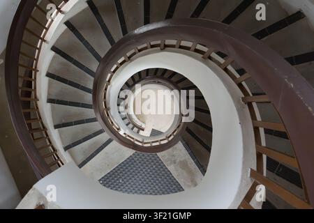 Circular staircase spirals downward in elegant curve. Dark treads contrast with white walls and brown railings. Central landing features a patterned t Stock Photo
