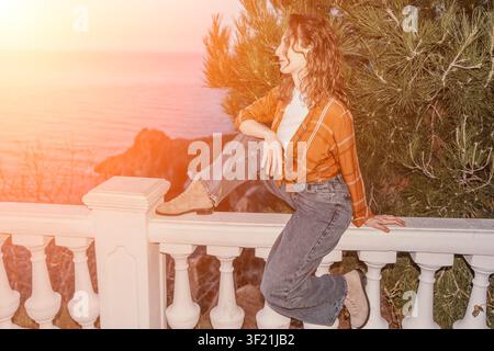 Woman balcony sunset sits on a white railing overlooking the ocean during golden hour for a peaceful vacation mood Stock Photo