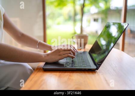 Horizontal close up of hands typing on laptop with coffee cup on wooden table in cozy home setting. Stock Photo