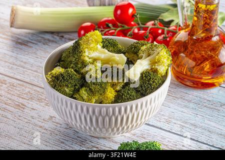 Steamed broccoli florets in textured white bowl surrounded by cherry tomatoes on vine glass oil bottle. Rustic wooden table background with natural li Stock Photo