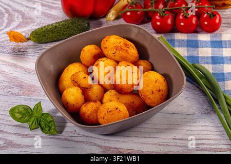 Golden roasted potatoes in gray bowl surrounded by cherry tomatoes cucumber green onions and basil on wooden surface with blue checkered cloth. Warm l Stock Photo