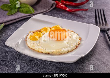 Closeup of fried chicken egg and three quail eggs on white square plate. Fresh basil leaves and red chili peppers in background. Black fork on grey st Stock Photo