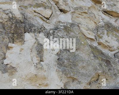 old stone wall made of uneven beige-toned limestone. The rough, textured surface reveals cracks, dark stains, gray spots, traces of weathering Stock Photo