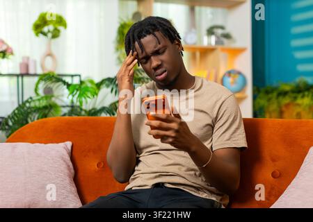 African American young man on home sofa holds smartphone upset after bad news message with stress disappointment. Black guy on home sofa sighs from online problem using phone in living room at home. Stock Photo