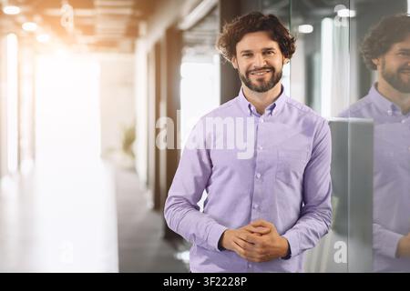 Man Stands Smiling in Office Corridor With Natural Light Shining Through Glass Walls Stock Photo