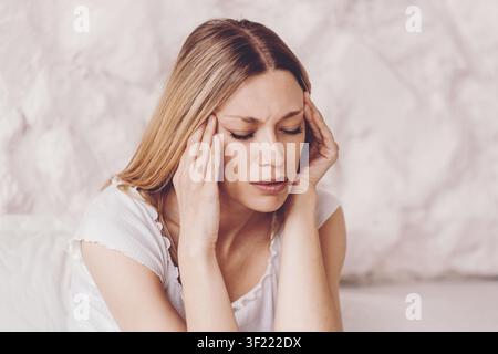Worried Young Woman With Long Hair Holding Head In Pain At Home, Healthcare Concept Stock Photo