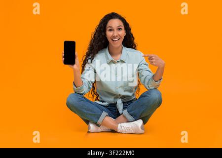 Woman Sitting on the Floor Shows Smartphone in Front of Bright Orange Background Stock Photo