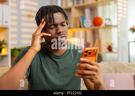 African American young man on home sofa looking at smartphone with sad face after bad news and online game loss. Black guy on home sofa feels upset while reading screen and reacting to online defeat. Stock Photo