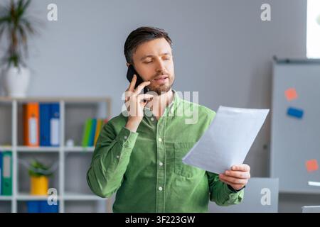 Focused Man In Green Shirt Talking On Phone While Reviewing Documents, Office Setting, Business Concept Stock Photo