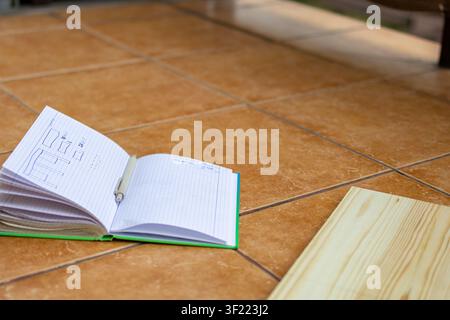 Open notebook with a hand drawn furniture sketch lies beside wooden boards on the floor before assembly begins. Concept of project planning, DIY furni Stock Photo