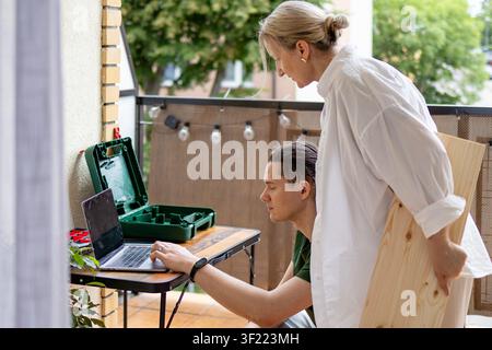 middle aged woman stands beside a man working with a laptop while planning a DIY furniture project on a balcony. Concept of online research, teamwork, Stock Photo