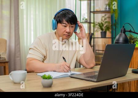 Asian man sitting at home table listens to podcast on laptop with headphones, nodding, writing notes in notebook. Chinese guy focuses, records insights, learning keenly through engaging audio session. Stock Photo