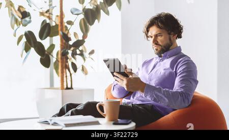 Man Reading on Tablet While Sitting in Bright Room With Plants and Coffee Stock Photo