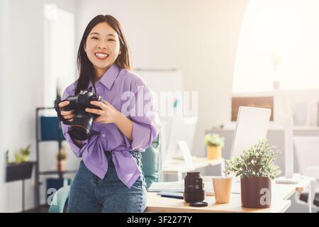 Smiling Young Asian Woman Holding Camera In Office, Photography Concept Stock Photo
