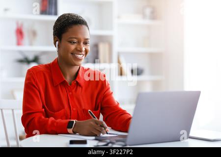 Woman Works on Laptop at Home While Taking Notes During Online Meeting in Bright Room Stock Photo