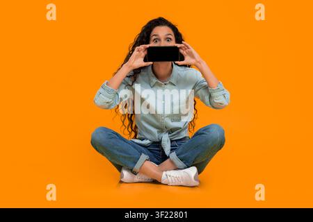 Young Woman Holds Smartphone in Front of Her Face While Sitting on Bright Background Stock Photo
