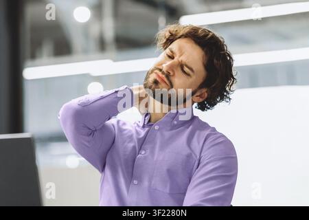 Man Sitting at Desk Rubbing Neck in Office During Work Hours While Wearing Purple Shirt Stock Photo