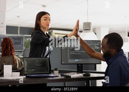 Male and female developers in modern office sharing high five after achieving app development goal. Happy colleagues celebrate successful programming project and teamwork in startup workplace. Stock Photo
