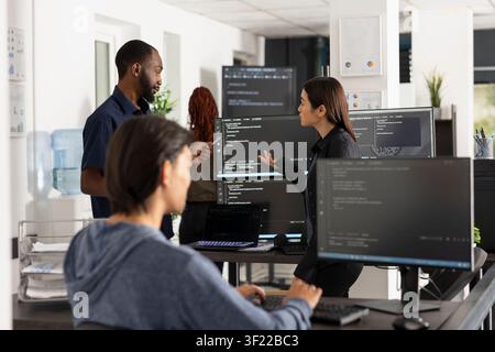Programmers collaborate at desktop monitor, comparing data to improve company website. Male and female coworkers review coding scripts together in office, highlighting teamwork in web development. Stock Photo