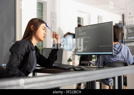 Female asian programmer works at desk, tapping pen while analyzing code on computer screen. Scene conveys concentration, innovation and digital workflow in modern software and web development. Stock Photo